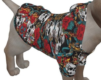May include: A small dog wearing a colorful shirt with a skull and rose pattern. The shirt features a black background with white skulls, red roses, and green leaves. The dog is light brown with a white background.