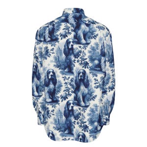 Puede incluir: Camisa de manga larga con un estampado de toile de Jouy azul y blanco. El patrón incluye ilustraciones de perros Cocker Spaniel, elementos florales y detalles arquitectónicos. La camisa está hecha de tela ligera.