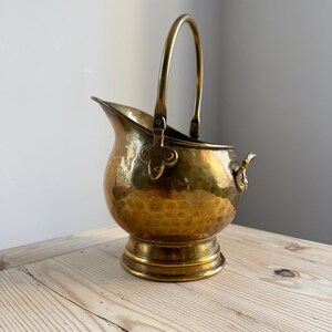 May include: A polished brass coal scuttle featuring a hammered texture on the rounded body, a wide pouring spout, a sturdy arched handle attached with clover-shaped metal mounts, and a circular pedestal base with a decorative side handle.