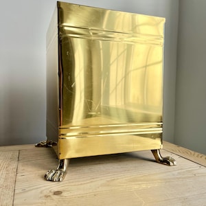 May include: A gold metal waste bin with a square shape and four lion paw feet. The bin has a shiny, polished finish.