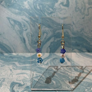 Puede incluir: Un par de pendientes de aro dorados con abalorios de flores azules, blancas y amarillas. Los pendientes se muestran en un soporte transparente sobre un fondo de mármol azul y blanco.