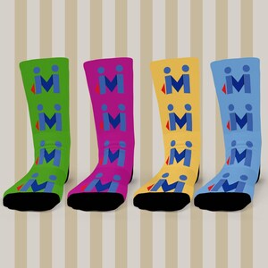 May include: Four brightly coloured socks, each in a different shade: green, purple, yellow, and blue. Each sock displays a repeating pattern of blue "M" shapes with a red accent. The socks have black toes and heels.