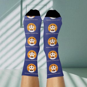 May include: Blue socks with a repeating pattern of a white cat face in an orange circle. The socks have black tops and the text "MASQUAS DOODLE SHOP" is printed on the socks.