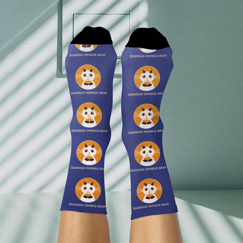 May include: Blue socks with a repeating pattern of a white cat face in an orange circle. The socks have black tops and the text "MASQUAS DOODLE SHOP" is printed on the socks.