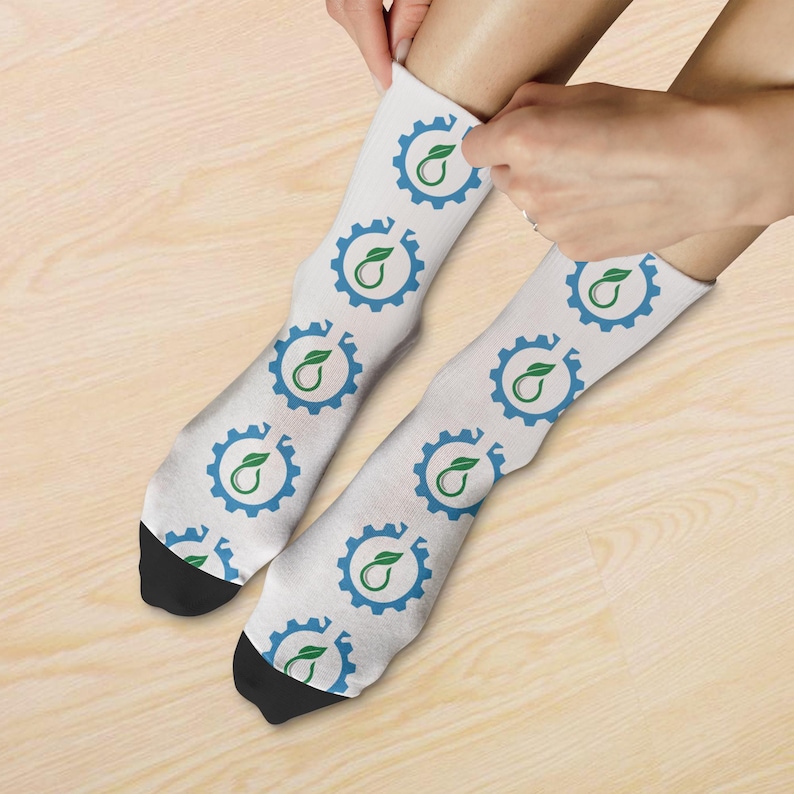 May include: White socks featuring a repeating pattern of a blue cogwheel and a green leaf design. The socks have black heels and toes. The socks are being put on.