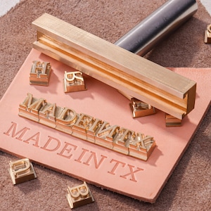 May include: A brass metal stamp holder with a T-shaped slot, accompanied by individual brass letter stamps spelling MADE IN TX. The stamps are arranged to form the text MADE IN TX on a piece of light brown leather, with additional loose letter stamps.
