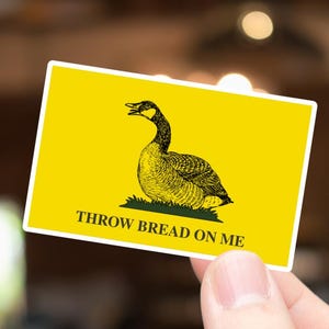 May include: A yellow sticker with a black goose illustration and the text "THROW BREAD ON ME". The goose is standing on a patch of green grass. The sticker is rectangular and held by a hand.