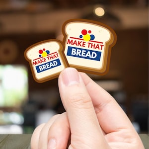 Może przedstawiać: Dwie naklejki w kształcie chleba z napisem "MAKE THAT BREAD" w kolorze czerwonym i niebieskim. Naklejki mają brązową sk&oacute;rkę i biały środek, z kolorowymi kropkami nad tekstem. Naklejki są trzymane w dłoni.