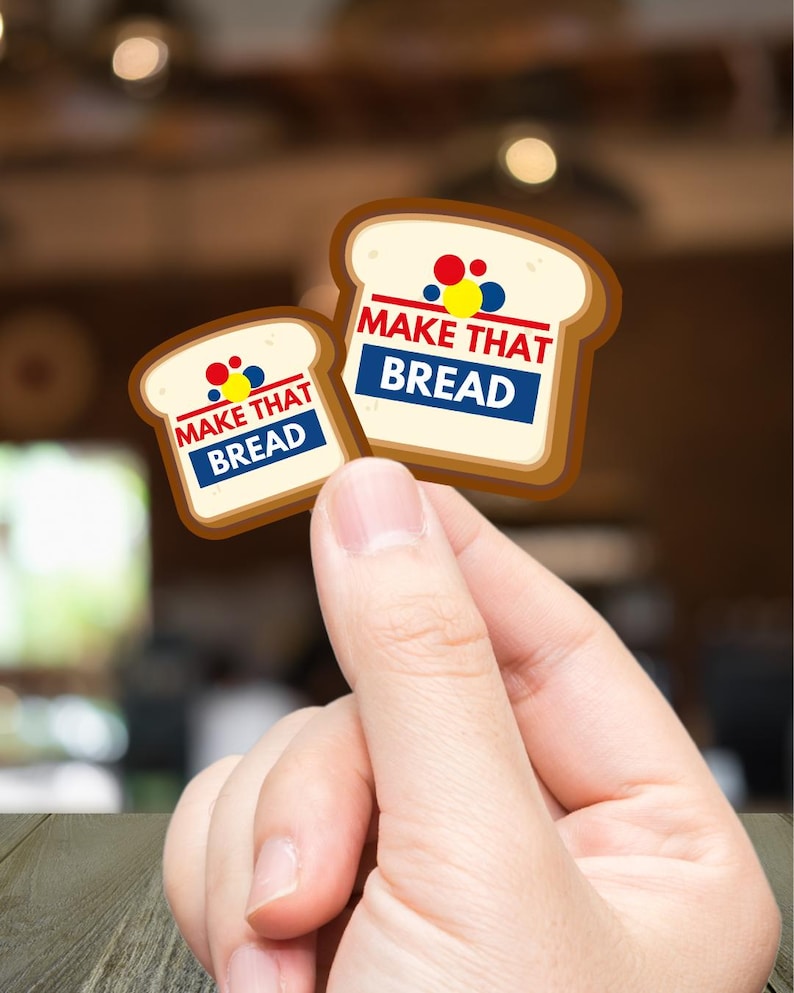 Może przedstawiać: Dwie naklejki w kształcie chleba z napisem "MAKE THAT BREAD" w kolorze czerwonym i niebieskim. Naklejki mają brązową sk&oacute;rkę i biały środek, z kolorowymi kropkami nad tekstem. Naklejki są trzymane w dłoni.
