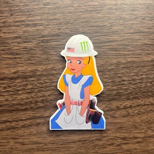 May include: A sticker featuring a cartoon image of Alice in Wonderland wearing a white hard hat with the American flag and a green Monster Energy logo. She is dressed in her classic blue and white dress, with a tool belt.