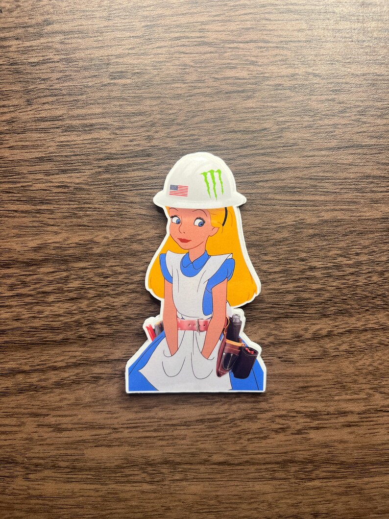 May include: A sticker featuring a cartoon image of Alice in Wonderland wearing a white hard hat with the American flag and a green Monster Energy logo. She is dressed in her classic blue and white dress, with a tool belt.
