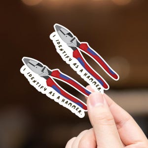 May include: Two stickers featuring a cartoon illustration of red and blue pliers with the text "I IDENTIFY AS A HAMMER." The pliers have silver blades and are held against a blurred background.