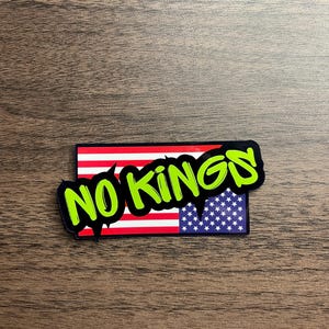 May include: A rectangular sticker featuring the American flag design with the words "NO KINGS" in bright green, dripping font. The sticker has a black border and is set against a wooden background.
