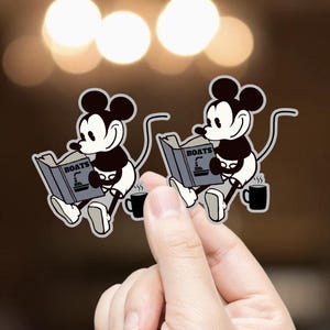 May include: Two stickers featuring a cartoon mouse reading a book titled "BOATS" with a ship illustration. The mouse is black and white, with a black coffee mug beside it. The stickers are held by a hand.