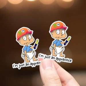 May include: Two stickers featuring a cartoon baby wearing an orange construction hat with a skull and green logo. The baby is in a blue shirt and diaper, holding a yellow and blue tool. Text on the sticker reads "I'm just an apprentice."