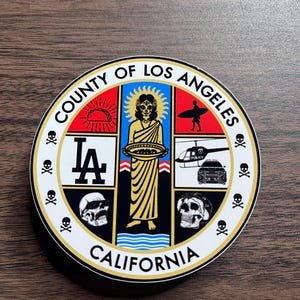 May include: A round sticker with a black and gold border. The sticker features a white background with the text "COUNTY OF LOS ANGELES CALIFORNIA" in a gold font. The center of the sticker features a black and white image of a coat of arms with a variety of symbols, including a skull, a taco, a surfboard, a helicopter, and a Los Angeles Dodgers logo.