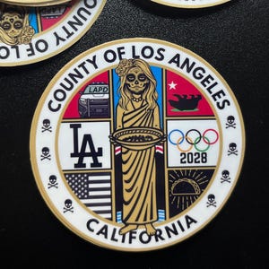 May include: A circular sticker featuring a stylized design with the text "COUNTY OF LOS ANGELES CALIFORNIA". The design includes a sugar skull, the LA Dodgers logo, the American flag, Olympic rings with "2028", and a taco.