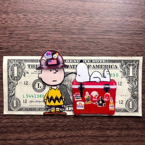 Może przedstawiać: Figurka Charliego Browna w kasku budowlanym z logo Taco Bell stoi obok czerwonej lod&oacute;wki z naklejkami. Snoopy wyleguje się na lod&oacute;wce, kt&oacute;ra leży na banknocie jednodolarowym.