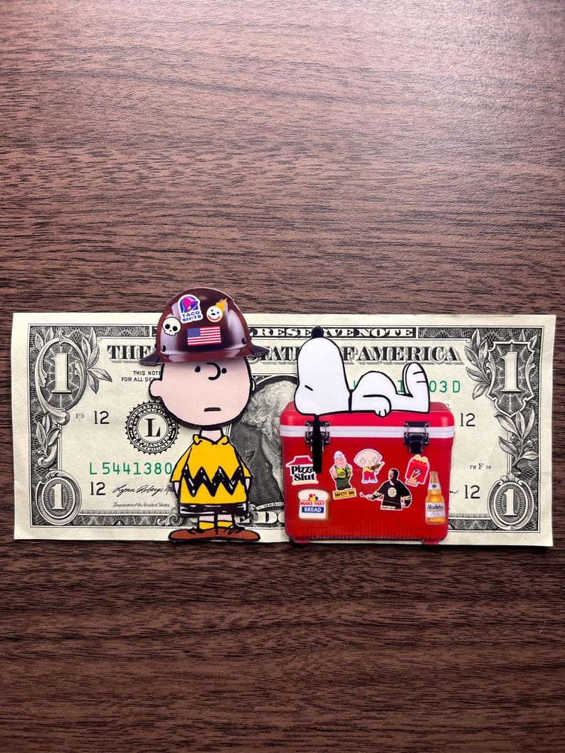 Może przedstawiać: Figurka Charliego Browna w kasku budowlanym z logo Taco Bell stoi obok czerwonej lod&oacute;wki z naklejkami. Snoopy wyleguje się na lod&oacute;wce, kt&oacute;ra leży na banknocie jednodolarowym.