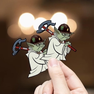 May include: Two cartoon Yoda figures wearing brown hard hats with a green Monster Energy logo and holding a hammer. The figures are green with white robes and are set against a blurred background.