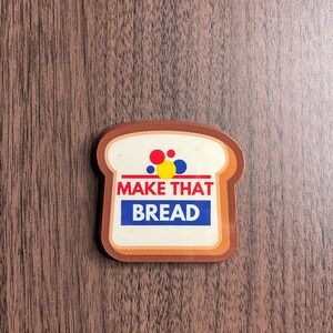 Może przedstawiać: Naklejka w kształcie chleba z napisem "MAKE THAT BREAD" w kolorze czerwonym i niebieskim. Naklejka ma biały środek z czerwonymi, ż&oacute;łtymi i niebieskimi kropkami. Obramowanie jest brązowe, a tło to usłojenie drewna.