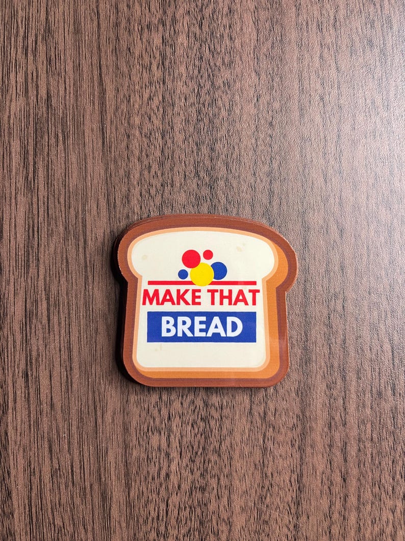 Może przedstawiać: Naklejka w kształcie chleba z napisem "MAKE THAT BREAD" w kolorze czerwonym i niebieskim. Naklejka ma biały środek z czerwonymi, ż&oacute;łtymi i niebieskimi kropkami. Obramowanie jest brązowe, a tło to usłojenie drewna.