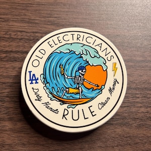 May include: A round sticker with a white background and a blue, green, and orange wave design. The sticker features a skeleton surfing on the wave with a beer in one hand. The text "Old Electricians Rule" is written in a circle around the image. The text "Dirty Hands Clean Money" is written in a circle around the image. The text "LA" is written in a circle around the image.