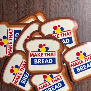 Może przedstawiać: Kolekcja naklejek w kształcie kromek chleba. Każda naklejka zawiera napis "MAKE THAT BREAD" w kolorze czerwonym i niebieskim, z grupą czerwonych, ż&oacute;łtych i niebieskich kropek. Naklejki znajdują się na brązowej drewnianej powierzchni.
