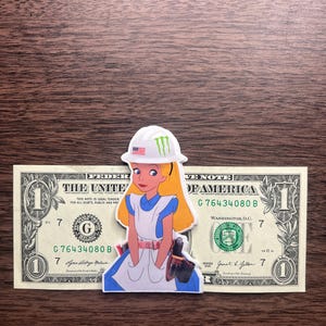 May include: A sticker of Alice in Wonderland wearing a white hard hat with an American flag and a green logo. The sticker is placed on top of a one-dollar bill, with the text "THE UNITED STATES OF AMERICA" visible.