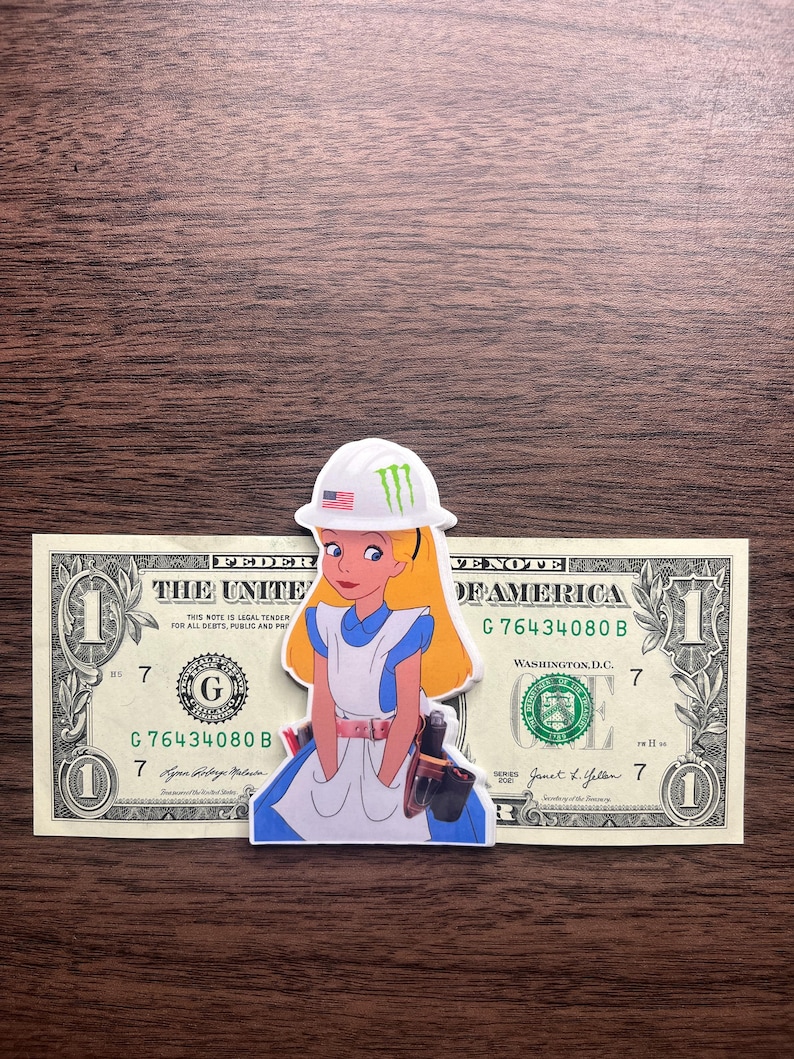 May include: A sticker of Alice in Wonderland wearing a white hard hat with an American flag and a green logo. The sticker is placed on top of a one-dollar bill, with the text "THE UNITED STATES OF AMERICA" visible.