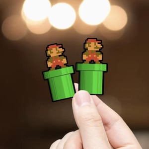 May include: Two pixelated Mario figures standing on green pipes. The figures are wearing red overalls, a red hat, and a brown mustache. The pipes are a bright green color. The background is blurred with warm, out-of-focus lights.