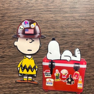 Może przedstawiać: Dwie naklejki z postaciami z kresk&oacute;wek. Jedna to Charlie Brown w brązowym kasku z naklejkami i ż&oacute;łtej koszuli. Druga to Snoopy na czerwonej lod&oacute;wce z naklejkami. Na lod&oacute;wce widnieje napis "Pizza Slut" i "Make That Bread".
