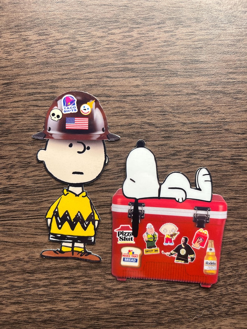 Może przedstawiać: Dwie naklejki z postaciami z kresk&oacute;wek. Jedna to Charlie Brown w brązowym kasku z naklejkami i ż&oacute;łtej koszuli. Druga to Snoopy na czerwonej lod&oacute;wce z naklejkami. Na lod&oacute;wce widnieje napis "Pizza Slut" i "Make That Bread".
