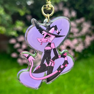 May include: A pink and black cartoon character with a witch's hat and a long tail. The character is wearing a black outfit with a sparkly design. The character is on a clear acrylic keychain.
