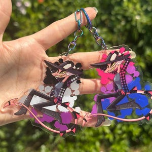 May include: Two acrylic keychains featuring a cartoon character with a black hat, plaid shirt, and devil tail. One keychain has pink and blue accents, the other has purple and gray accents. Both are held up by a hand.