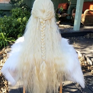 May include: A long, light blonde wig with a center braid and loose, wavy strands. The wig is adorned with small gold beads and is paired with a white faux fur cape, creating a fantasy or cosplay aesthetic.