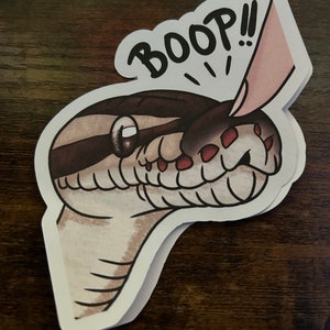 May include: A sticker featuring a cartoon snake head with brown and tan markings. The word "BOOP!!" is written above the snake's head, with a pink finger booping the snake's head. The sticker has a white border.