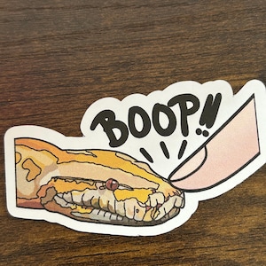 May include: A sticker of a yellow and brown snake with a red eye, being "booped" by a finger. The sticker has the word "BOOP!" written above the snake.