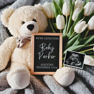 Puede incluir: Un oso de peluche blanco se sienta en una manta gris con un letrero de madera que dice "We're expecting Baby Parker Arriving December 2024". Hay flores blancas en el fondo.