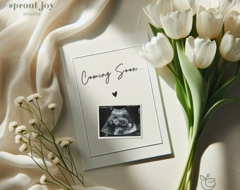May include: A white frame with a black and white ultrasound image and the text "Coming Soon..." with a small heart symbol. The frame is on a white fabric background with a bouquet of white tulips and a sprig of white flowers.