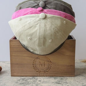 Könnte beinhalten: Ein Hutständer aus Holz mit einer personalisierten Inschrift, die "MIKE" lautet, und einem Lorbeerkranz mit dem Initial "M" in der Mitte. Der Ständer hält drei Baseballkappen, eine beige, eine rosa und eine braune.