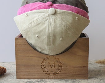 May include: Wooden hat rack with a personalized inscription that reads "MIKE" and a laurel wreath with the initial "M" in the center. The rack is holding three baseball caps, one beige, one pink, and one brown.