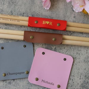 May include: Drumsticks with a red and brown leather holder. The red holder has gold lettering and a heart symbol. Two square coasters are visible, one grey with "Champagne" and one pink with "Michaela" printed on them.