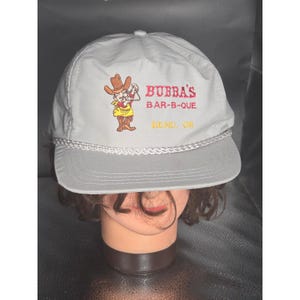 Op de afbeelding: Een lichtgrijze baseballpet met een geborduurde afbeelding van een cartooncowboy die een triangel vasthoudt. De tekst BUBBA'S BAR-B-QUE en BEND, OR is in rood en geel geborduurd. Een wit gevlochten koord siert de platte klep.