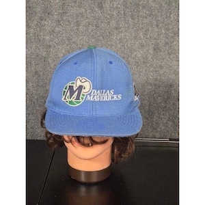 Puede incluir: Gorra de béisbol azul claro con el logotipo de los Dallas Mavericks. El logotipo presenta una "M" estilizada con una pelota de baloncesto y las palabras "DALLAS MAVERICKS" en blanco. La gorra tiene una visera verde.