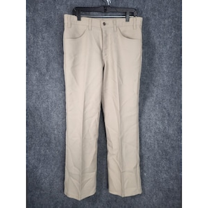 Puede incluir: Pantalones color beige con cierre de botón y cremallera, con bolsillos delanteros y traseros. Los pantalones cuelgan de una percha negra sobre un fondo gris. Los pantalones tienen un diseño clásico de pierna recta.