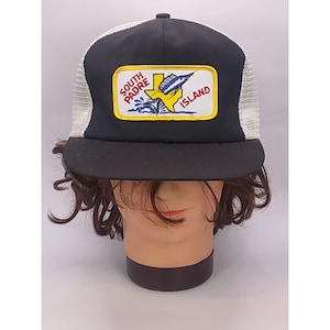 May include: A black and white trucker cap featuring a rectangular patch on the front. The patch displays a yellow outline of Texas, a blue marlin fish, and the text South Padre Island in red and blue lettering against a white background.