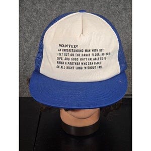Könnte beinhalten: Blau-weiße Trucker-Cap mit dem Text "WANTED: AN UNDERSTANDING MAN WITH HOT FEET OUT ON THE DANCE FLOOR. NO HAIR LIPS, AND GOOD RHYTHM, ABLE TO FURNISH A PARTNER WHO CAN FHOLICK ALL NIGHT LONG WITHOUT FAIL."