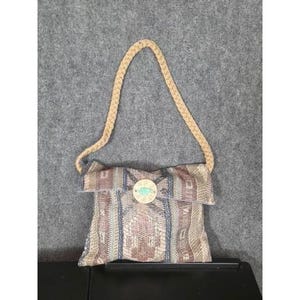 以下が含まれることがあります： ベージュの編み込みストラップが付いた、織り生地のハンドバッグ。バッグは、茶色、青、ピンクの色合いの幾何学模様が特徴です。ターコイズのデザインの丸い木製ボタンがフラップを固定しています。