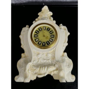 May include: Ivory-colored mantel clock with a gold-toned clock face featuring black Roman numerals. The clock has an ornate, scrolled design and a decorative base. The clock face is approximately 4 inches in diameter.